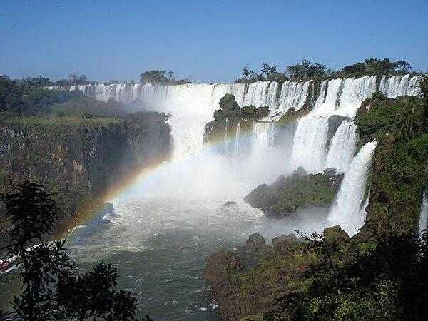 A rainbow appears in the mist of Iguazu Falls. The falls are part of a nearly virgin jungle ecosystem surrounded by national parks on both the Argentine and the Brazilian sides of the cascades. The Iguazu River begins in Parana state of Brazil, then crosses a 1,200-km (750 mi) plateau before reaching a series of faults forming the falls.
