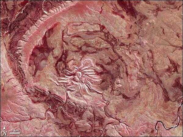 Over the Kimberley Region of northern Western Australia, satellite sensors and airplane passengers alike can see a giant arachnid sprawling over the arid landscape. This spider is not just big, it is old. This prehistoric monster crawls out of the past as if to remind us of the destructive power of the cosmos. In this false-color satellite image Spider Crater and the surrounding arid landscape appear in varying shades of crimson. Water appears blue-black, namely in the meandering river near the bottom edge of the image. Vegetation appears in shades of red. While vegetation looks sparse throughout the area, the intense red dots along the river indicate fairly lush - if intermittent - vegetation lining the riverbanks.

Strongly deformed layers of sedimentary rock give evidence of that the structure was formed by extraterrestrial trauma. Spider Crater rests in a depression some 13 by 11 km (8 by 7 mi) across. Meteorite craters often have central areas of uplift, and Spider Crater fits this pattern, with a central dome roughly 500 m (1,640 ft) in diameter. Radiating from this central dome are features unusual in impact craters in general, but important in giving this crater its nickname. Overlapping beds of tough sandstone that have weathered the elements far better than the surrounding rocks form the spider&apos;s &quot;legs.&quot; So while Spider Crater sits in a depression and has a central uplift area characteristic of impact craters, it shows extreme differences in erosion, giving it a unique appearance. The age of Spider Crater is uncertain, but its formation has been estimated to fall between 900 and 600 million years ago. Image courtesy of NASA.