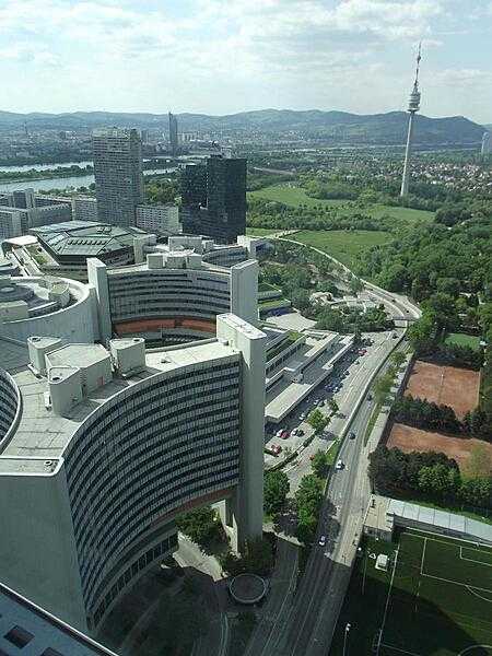 A bird&apos;s-eye view of two of the six Y-shaped office buildings, as well as the central cylindrical conference center, that make up part the United Nations Office complex in Vienna. In the distance is the 252 m (827 ft) Donauturn (Danube Tower) the tallest structure in Austria. In addition to its use as a communication tower, it also houses two revolving retaurants and has an observation deck.