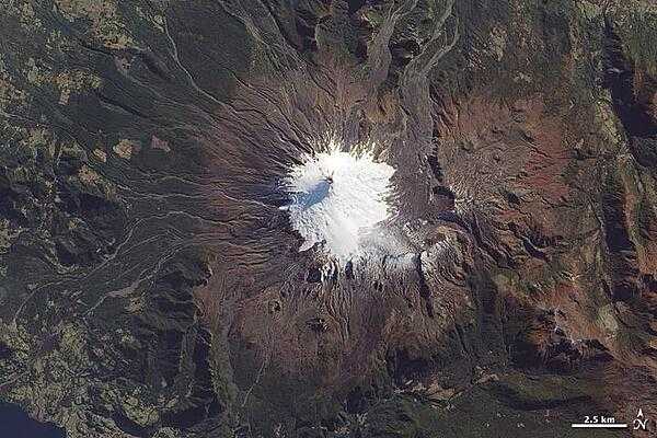 Near the southern tip of South America, a trio of volcanoes lines up perpendicular to the Andes Mountains. The most active is the westernmost, Volcan Villarrica, pictured in this photo-like satellite image. The 2,582-m (9,357-ft) stratovolcano is mantled by a 30-sq km (10-sq mi) glacier field, most of it amassed south and east of the summit in a basin made by a caldera depression. To the east and northeast, the glacier is covered by ash and other volcanic debris, giving it a rumpled, brown look. The western slopes are streaked with innumerable gray-brown gullies, the paths of lava and mudflows (lahars). Beyond the reach of ash and debris deposits, the volcano is surrounded by forests; the area is a national park. The largest recent eruption occurred in the early 1970s; lava flows melted glaciers and generated lahars that spread at speeds of 30-40 km per hour (20-30 mph). Image courtesy of NASA.