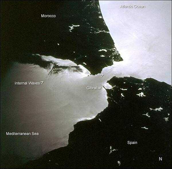 This space shuttle photo was taken near sunset. Two packets of tidally-generated internal waves are highlighted by sunglint off the surface waters in the Strait of Gibraltar. The older packet (labeled) contains at least 14 waves, which can be counted like tree rings. A younger group is forming near the middle of the strait (marked by the carat south of Gibraltar). The waves are generated as a diurnal tidal pulse flows over the shallow Camarinal Sill at Gibraltar. The waves flow eastward and refract around coastal features; they can be traced for as much as 150 km. Image credit: NASA.