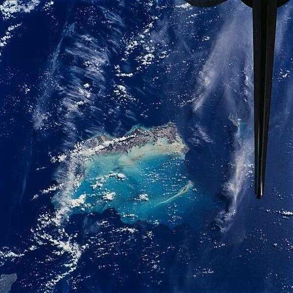 The Turks and Caicos Islands as seen from the space shuttle. In this view, the extensive shallow water areas of Caicos Bank (turquoise) dominate to the south of the Caicos Islands. Caicos Bank covers an area of 7,680 sq km (2,965 sq mi). The coral reefs of Caicos are primarily along the north deep water edge of the islands, and in a barrier to the south of the bank. East of Caicos Bank, near the tail of the shuttle, is the island of Grand Turk, part of the much smaller Turks Bank. The channel that runs between the two banks is more than 2,200 m (1.4 mi) deep. Image courtesy of NASA.