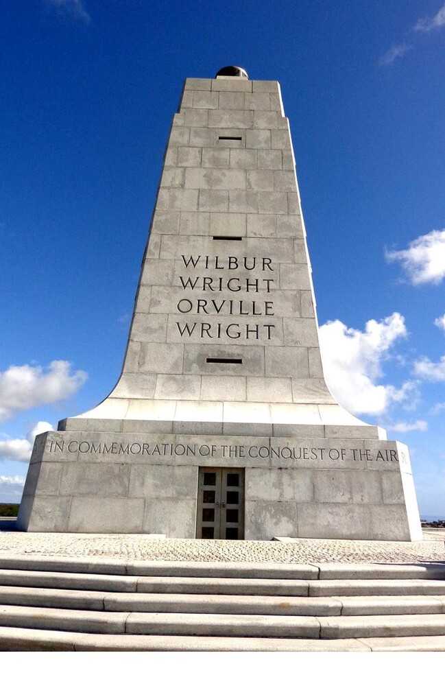 On 17 December 1903, Orville Wright, working with his brother Wilbur, flew the first successful manned, powered, controlled, heavier-than-air flight at Kitty Hawk, North Carolina. The Wright Brothers National Memorial, located in Kill Devil Hills near Kitty Hawk, is on the spot where the brothers tested their flying machines for three years before that successful 1903 flight. The two designed and built their gliders and aircraft at their home in Dayton, Ohio and tested them in privacy in North Carolina.  The monument, dedicated in December 1932, is an 18.29 m (60 ft) granite monument atop 27 m (90ft) tall Kill Devil Hill. On each side is a wing-like design, a lit beacon at the top, and an inscription “In commemoration of the conquest of the air by the brothers Wilbur and Orville Wright. Conceived by genius. Achieved by dauntless resolution and unconquerable faith.”